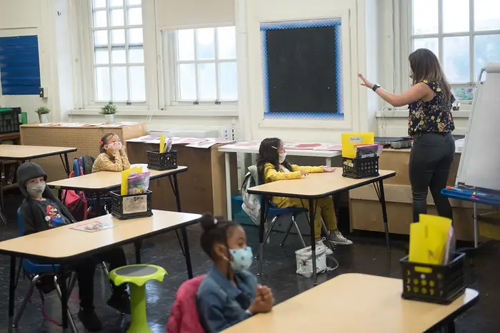 A teacher stands in front of a classroom where children are sitting at desks largely apart.